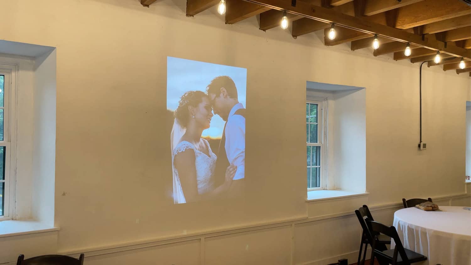 A large projection of a bride and groom displayed on a white wall in a room with wooden beams and tables set for an event.